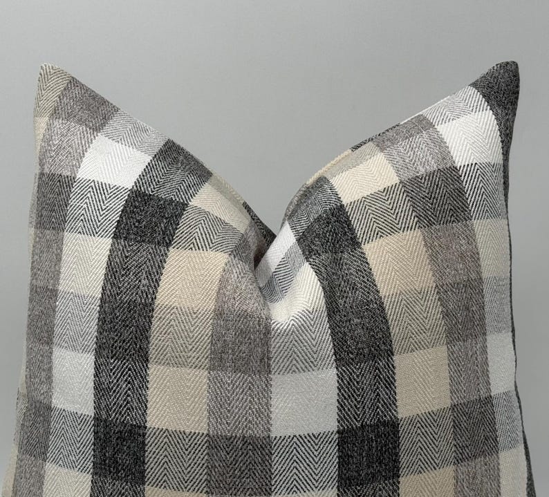 May include: A decorative pillow with a large, checkered pattern in shades of gray, white, and beige. The pillow has a soft, textured appearance, with a classic, neutral design. The pillow is a square shape.