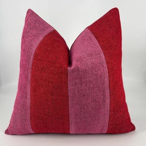 May include: A square throw pillow featuring a color-blocked design with vertical panels of deep red and bright pink fabric. The textile has a visible woven texture throughout, creating a consistent appearance across the front of the cushion.