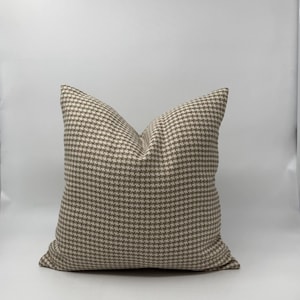 May include: A square decorative pillow with a houndstooth pattern in shades of beige and brown. The pillow is made of a textured fabric and is designed for home decor.