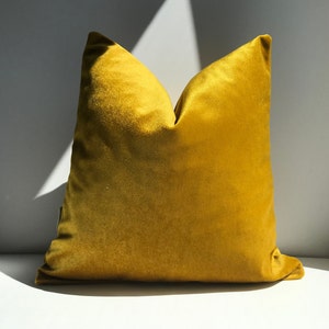 May include: A mustard yellow velvet pillow with a square shape. The pillow is sitting on a white surface.