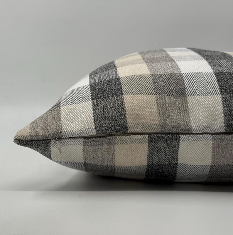 May include: A decorative pillow with a plaid pattern in shades of gray, beige, and white. The pillow has a herringbone weave and a zipper closure. The pillow is a rectangular shape.