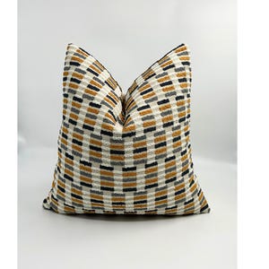 May include: A decorative pillow with a textured, geometric pattern. The pillow features a repeating design of mustard yellow, black, gray, and off-white rectangles. The pillow is square and appears to be made of a soft fabric.