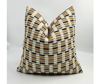 May include: A decorative pillow with a textured, geometric pattern. The pillow features a repeating design of mustard yellow, black, gray, and off-white rectangles. The pillow is square and appears to be made of a soft fabric.