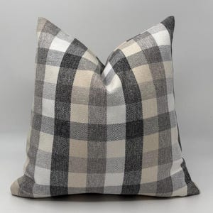 May include: A square decorative pillow with a large plaid pattern in shades of gray, white, and beige. The pillow has a soft, textured appearance, suitable for home decor. The pillow's dimensions are not visible.