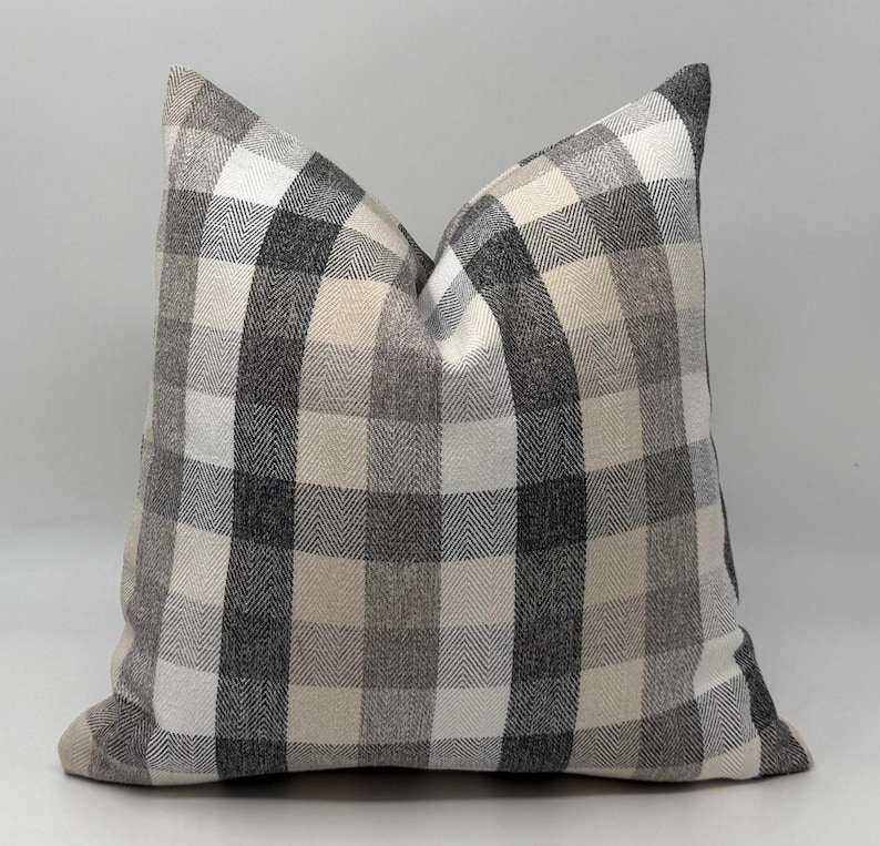 May include: A square decorative pillow with a large plaid pattern in shades of gray, white, and beige. The pillow has a soft, textured appearance, suitable for home decor. The pillow's dimensions are not visible.