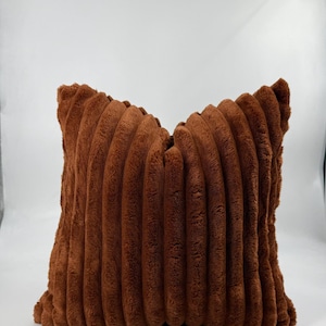 May include: A square, brown decorative pillow with a corduroy-like texture. The pillow has a soft, plush appearance and is designed for home decor. The color is a warm, earthy tone, suitable for various interior styles.
