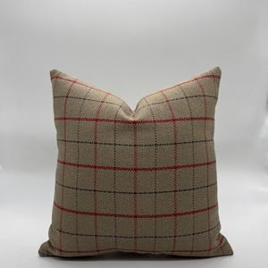 May include: A square decorative pillow with a beige background and a plaid pattern in red and black. The pillow is made of a textured fabric and is designed for home decor.