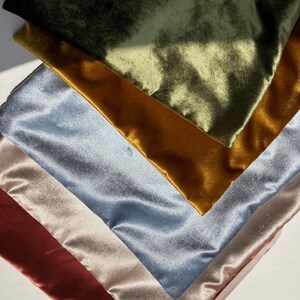 May include: A stack of five fabric swatches in different colors and textures. The top swatch is a dark green velvet, followed by a mustard yellow velvet, a light blue velvet, a light beige satin, and a burgundy satin.