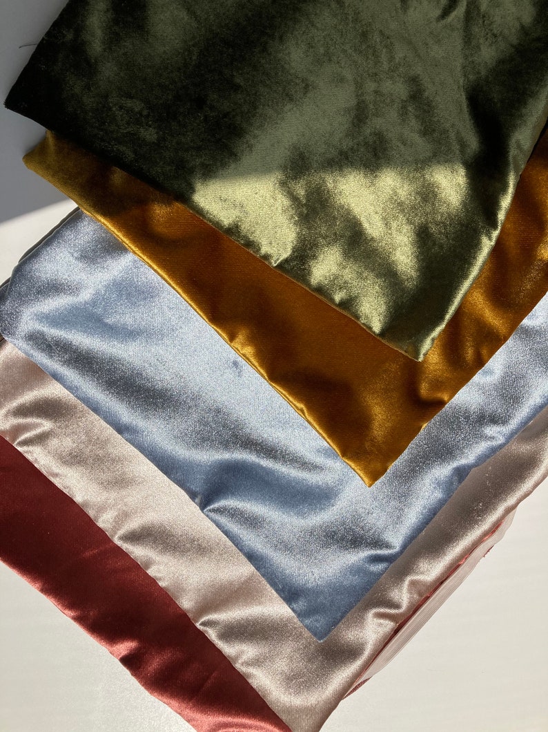 May include: A stack of five fabric swatches in different colors and textures. The top swatch is a dark green velvet, followed by a mustard yellow velvet, a light blue velvet, a light beige satin, and a burgundy satin.