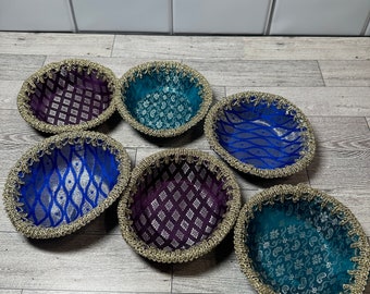 Puede incluir: Seis cuencos decorativos pequeños con ribete dorado. Los cuencos están forrados con tela en varios patrones y colores, incluyendo morado, azul y turquesa.