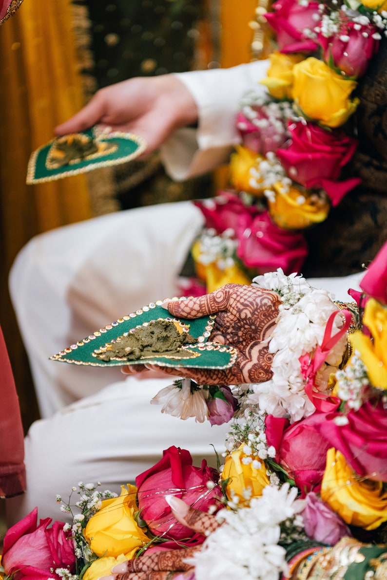 May include: A close-up of a hand holding a green heart-shaped tray with a brown paste. The hand is decorated with henna and the tray is adorned with gold glitter. The background is blurred and features a bouquet of pink and yellow roses.