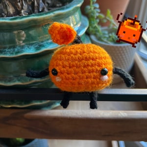 May include: A handmade orange crochet creature with black arms and legs, and a small orange top. The creature has black eyes and blush. The image also includes a pixelated orange character.