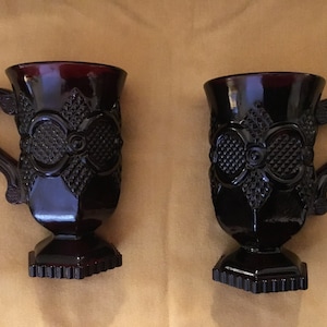 Puede incluir: Dos tazas de vidrio rojo oscuro con un patrón decorativo y un asa. Las tazas tienen una base con pie.