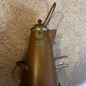 May include: A copper-colored vintage tea kettle with a handle and spout. The kettle has a lid with a small, dark knob. A small tag with the text "HAND CRAFTED COPPERWARE" and "DOURO" is visible.
