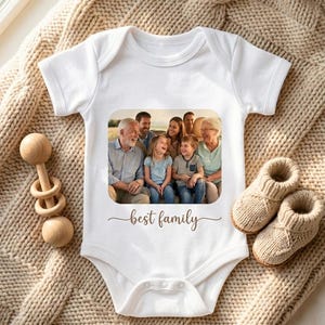 May include: A white short-sleeved baby bodysuit featuring a rectangular colour photograph of a multi-generational family and the cursive text "best family" printed below the image. The garment is displayed alongside a wooden rattle and booties.