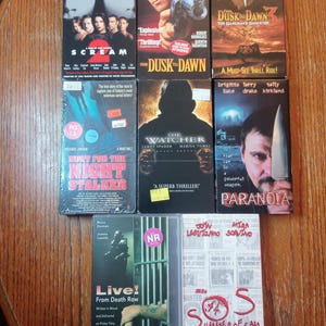 May include: A collection of VHS tapes featuring movie titles like "Scream 2", "From Dusk Till Dawn", and "Summer of Sam". The tapes are arranged on a wooden surface, showcasing various horror and thriller film covers.