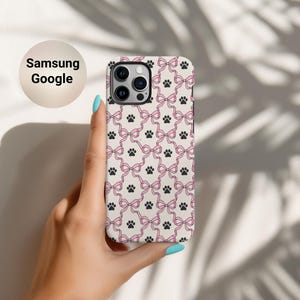 Op de afbeelding: Een telefoonhoesje met een herhaald patroon van roze strikken en zwarte pootafdrukken op een crèmekleurige achtergrond. De hoes wordt vastgehouden in een hand met blauwe nagellak. De tekst "Samsung Google" staat in een cirkel linksboven.