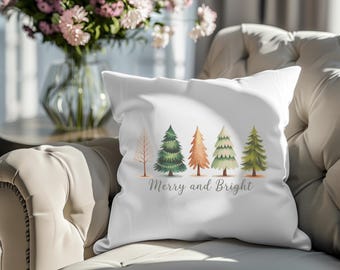 Puede incluir: Cojín cuadrado blanco con un diseño de acuarela de cinco árboles de Navidad de varios colores. El texto "Merry and Bright" está escrito debajo. El cojín está sobre un sofá beige.