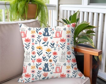 May include: A decorative throw pillow with a floral and cat pattern. The pillow features a cream background with pink, orange, and blue flowers, and gray, pink, and blue cat illustrations. The pillow is placed on a beige outdoor sofa.