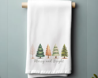 May include: White kitchen towel with a watercolor design of five Christmas trees in varying shades of green, orange, and brown. The text "Merry and Bright" is printed below the trees. The towel is hanging on a wooden rod.