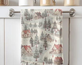 May include: A kitchen towel with a winter village scene. The design features houses with red roofs and gray trees on a cream background. The towel is hanging on a silver bar in front of a white cabinet with jars on a shelf.