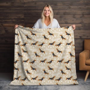 Peut inclure: Couverture beige avec un motif répété de chiens beagle, d'empreintes de pattes et le mot "Beagle" en blanc. La couverture est faite d'une matière douce et convient à une utilisation dans un salon ou une chambre.