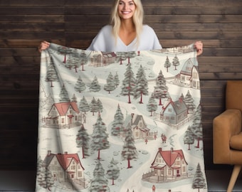 May include: A cozy blanket featuring a winter village scene with houses, trees, and winding roads. The blanket has a cream-colored background with red, gray, and brown details. The blanket is held up by a person.