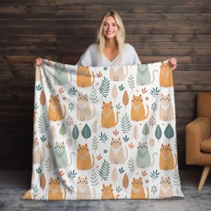 Peut inclure: Une couverture blanche avec un motif répété de chats de dessin animé dans les tons orange, beige et vert, ornée de feuilles et d'éléments floraux. La couverture est tenue par une personne.