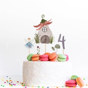 May include: A white frosted cake decorated with colorful macarons and fairy-themed toppers. The toppers include a fairy house, a fairy, mushrooms, flowers, and the number 4. Sprinkles are scattered around the base of the cake.