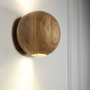 Op de afbeelding: Een houten wandlamp met een ronde, lichtbruine kap. Het licht is aan en werpt een lichtstraal op de muur.