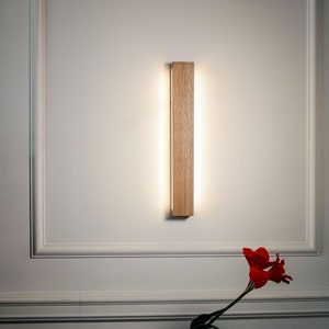 May include: A wooden wall sconce with a warm white light illuminates a white wall. A single red flower is in a dark blue vase on a glass table.