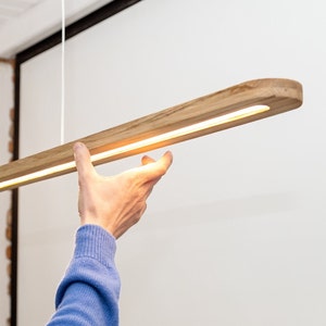 Może przedstawiać: Drewniana lampa z ciepłym białym paskiem LED. Lampa jest zawieszona na suficie i jest trzymana przez rękę.