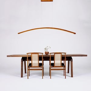 May include: A wooden dining table with two chairs and a curved wooden light fixture hanging above. The table is made of dark wood and has a natural finish. The chairs are also made of wood and have a woven seat.