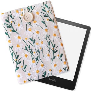 K&ouml;nnte beinhalten: Eine wei&szlig;e und gr&uuml;ne, floral gemusterte Kindle E-Reader-H&uuml;lle mit einem wei&szlig;en Knopf. Die H&uuml;lle liegt auf einem schwarzen Kindle E-Reader mit dem Wort "kindle" auf der Unterseite.
