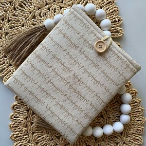 May include: A beige fabric pouch with a brown tassel and a white button closure. The pouch is decorated with brown script writing and has a white background. The pouch is sitting on a woven straw mat with white beads.