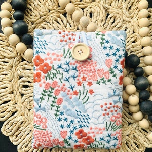 May include: A white fabric phone case with a floral embroidery design in shades of pink, blue, and green. The case has a button closure and a white drawstring.