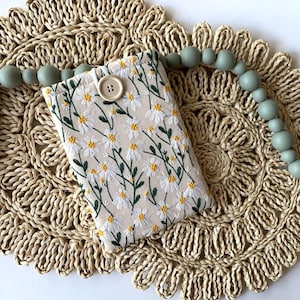 K&ouml;nnte beinhalten: Eine kleine wei&szlig;e Stofftasche mit Blumenmuster und einem Knopfverschluss. Die Tasche liegt auf einem geflochtenen Stroh-Untersetzer mit einer Schnur aus hellgr&uuml;nen Perlen.