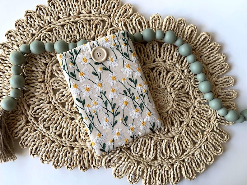 K&ouml;nnte beinhalten: Eine kleine wei&szlig;e Stofftasche mit Blumenmuster und einem Knopfverschluss. Die Tasche liegt auf einem geflochtenen Stroh-Untersetzer mit einer Schnur aus hellgr&uuml;nen Perlen.