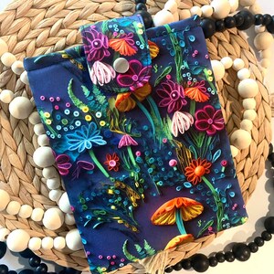 May include: A dark blue phone case with a colorful floral and mushroom design. The case has a button closure and a tassel.