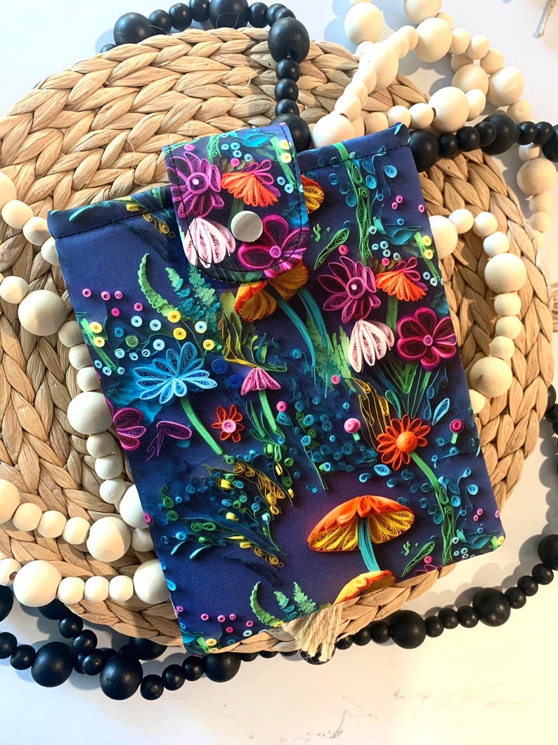 May include: A dark blue phone case with a colorful floral and mushroom design. The case has a button closure and a tassel.
