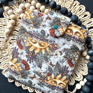 May include: A beige fabric pouch with a floral pattern featuring suns, mushrooms, and butterflies. The pouch has a green button closure.