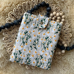 K&ouml;nnte beinhalten: Eine beige Stoffh&uuml;lle f&uuml;r ein Handy mit einem gestickten Muster aus wei&szlig;en G&auml;nsebl&uuml;mchen. Die H&uuml;lle hat eine kleine Klappe mit einem Knopfverschluss.