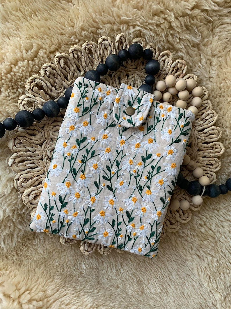 K&ouml;nnte beinhalten: Eine beige Stoffh&uuml;lle f&uuml;r ein Handy mit einem gestickten Muster aus wei&szlig;en G&auml;nsebl&uuml;mchen. Die H&uuml;lle hat eine kleine Klappe mit einem Knopfverschluss.