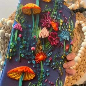 May include: A dark blue clutch bag with a colorful floral and mushroom design. The flowers are made of paper and are in shades of pink, orange, yellow, and green. The mushrooms are orange and yellow.