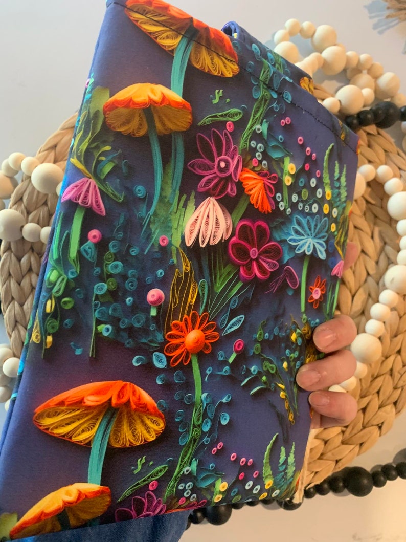 May include: A dark blue clutch bag with a colorful floral and mushroom design. The flowers are made of paper and are in shades of pink, orange, yellow, and green. The mushrooms are orange and yellow.