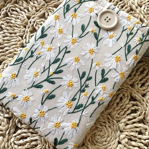 K&ouml;nnte beinhalten: Eine wei&szlig;e Stofftasche mit einem Blumenmuster aus gestickten wei&szlig;en G&auml;nsebl&uuml;mchen und gr&uuml;nen Stielen. Die Tasche hat einen braunen Knopfverschluss.