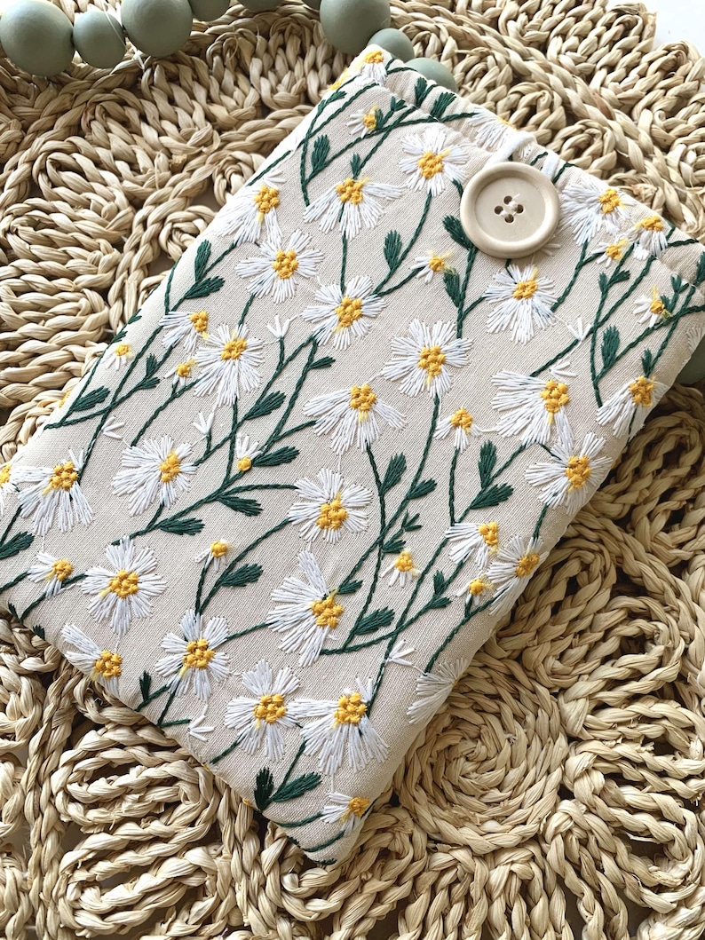 K&ouml;nnte beinhalten: Eine wei&szlig;e Stofftasche mit einem Blumenmuster aus gestickten wei&szlig;en G&auml;nsebl&uuml;mchen und gr&uuml;nen Stielen. Die Tasche hat einen braunen Knopfverschluss.