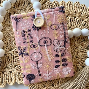 May include: A pink fabric book cover with embroidered floral designs in white, gold, and navy blue. The cover has a wooden button closure and a white loop. The background includes a woven mat and decorative wooden beads.