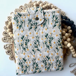 K&ouml;nnte beinhalten: Eine beige Stofftasche mit einem gestickten Muster aus wei&szlig;en G&auml;nsebl&uuml;mchen. Die Tasche hat einen Klappverschluss mit einem Knopf.