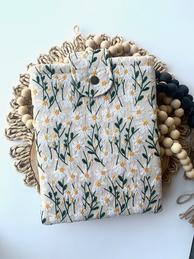 K&ouml;nnte beinhalten: Eine beige Stofftasche mit einem gestickten Muster aus wei&szlig;en G&auml;nsebl&uuml;mchen. Die Tasche hat einen Klappverschluss mit einem Knopf.